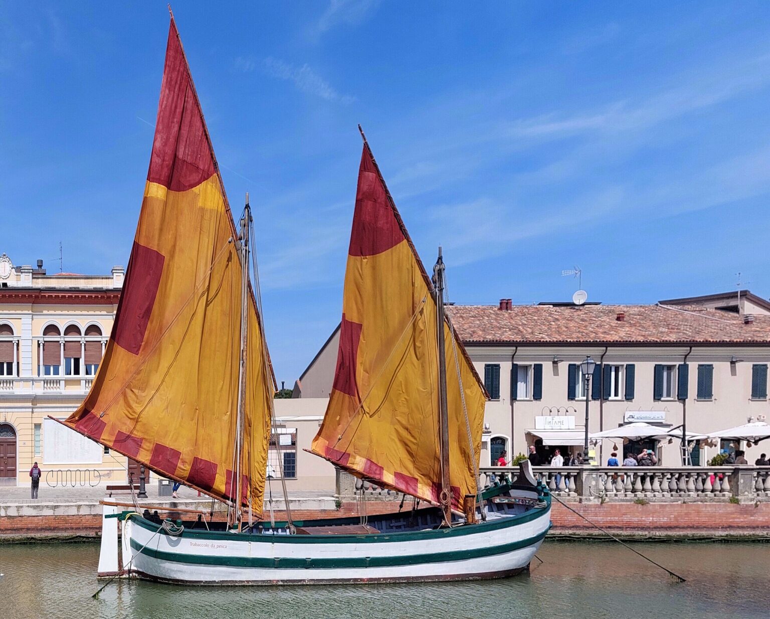 A large wooden cargo boat, with two masts and two yellow/red lateen (triangular) sails. In the background old houses and a quayside.