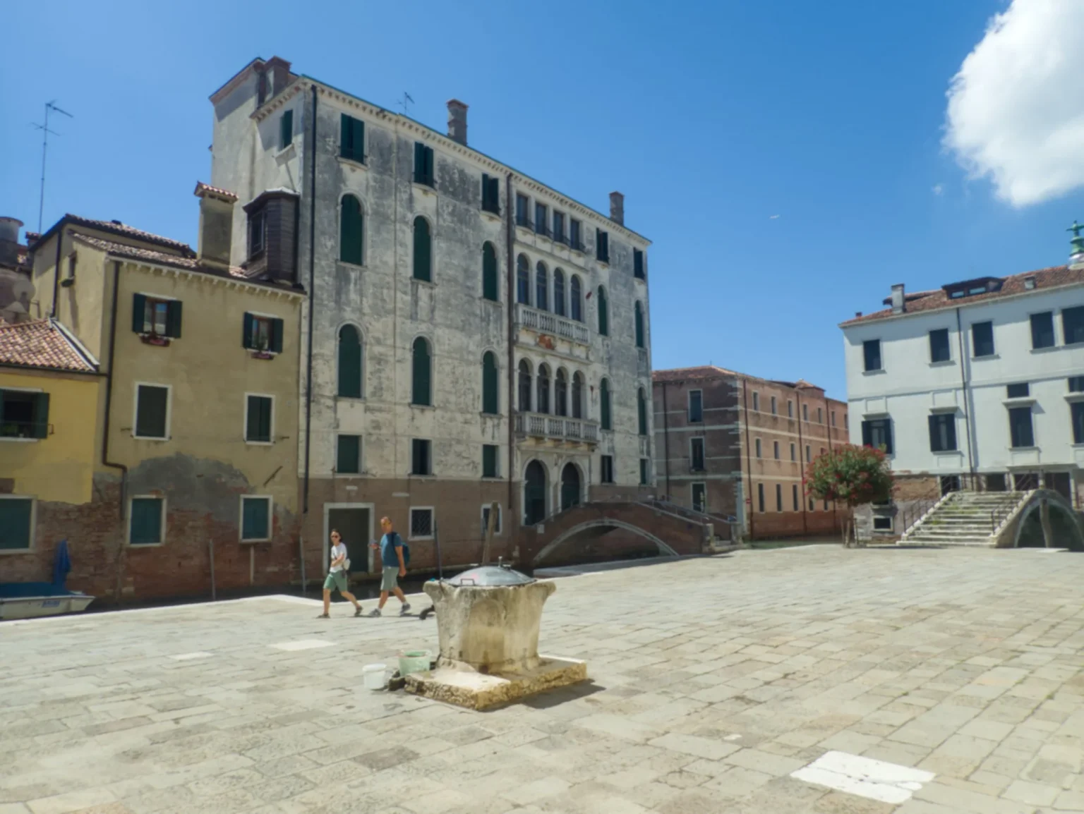 In the foreground an almost empty square with a well-head in the middle. There are only two persons walking across the otherwise deserted space. Behind the square, connected by a bridge, stands a grimey off-white Venetian palace of four or five storeys, noticeably taller than the surrounding houses. All the windows have closed shutters. To the left, a smaller three storey yellow house, and to the right, a well-kept white building of three storeys, also with a bridge in front.