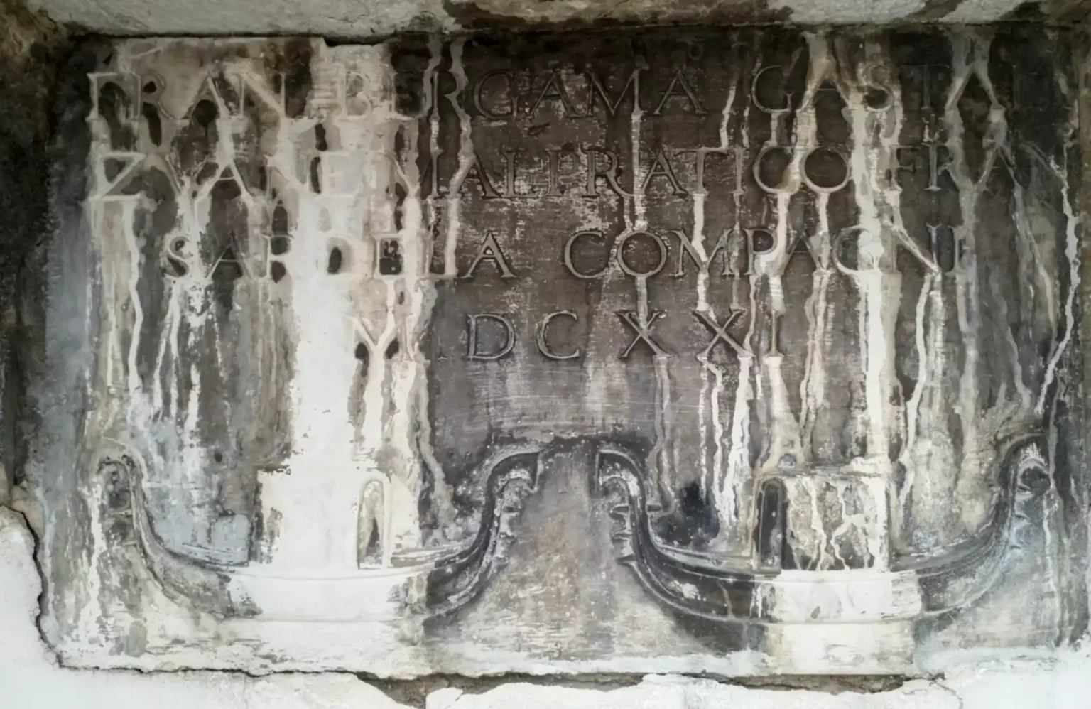 A stone plaque, mostly dark brown but with yellow vertical bands caused by flowing water. It has a four line inscription in all capital letters, which is difficult to read due to the discolouring. Below, reliefs of two banana-shaped boats, each with a central cabin, and ornate decorations front and back.