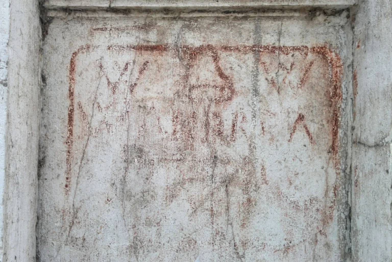 A shallow niche in a yellowish and dirty stone wall, with markings in red painted on it. The markings are within a rectangular frame with rounded corners, in the same colour. The first line has a W, a simple drawing of a pointed hat, and another W. Second line has a W and the letters A N D R E A, only partially readable. The last two lines are not readable.
