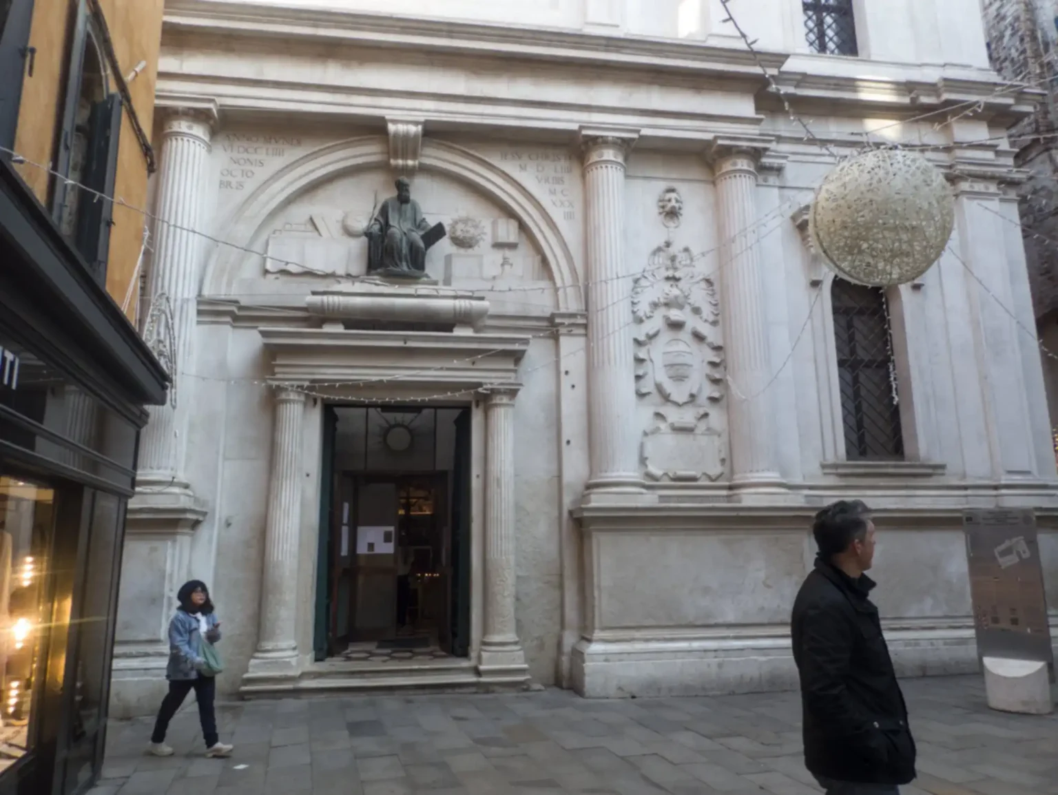 The lower order of the façade of San Zulian as it appears to the casual passer-by, in the rather cramped space. The main doorway to the church is surmounted by a half-circular space, with the statue of the sitting Ravenna surrounded by his books and symbols. Around the doorway is a kind of triumphal arch, with the coat of arms of Ravenna, and the Greek inscription on the right (visible) and the Hebrew (out of frame) to the left.